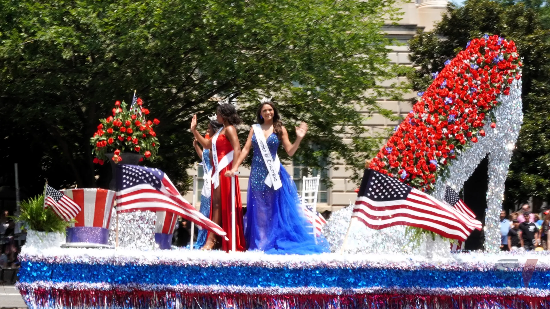 Two contestants in vibrant dresses waving on a float during a parade.