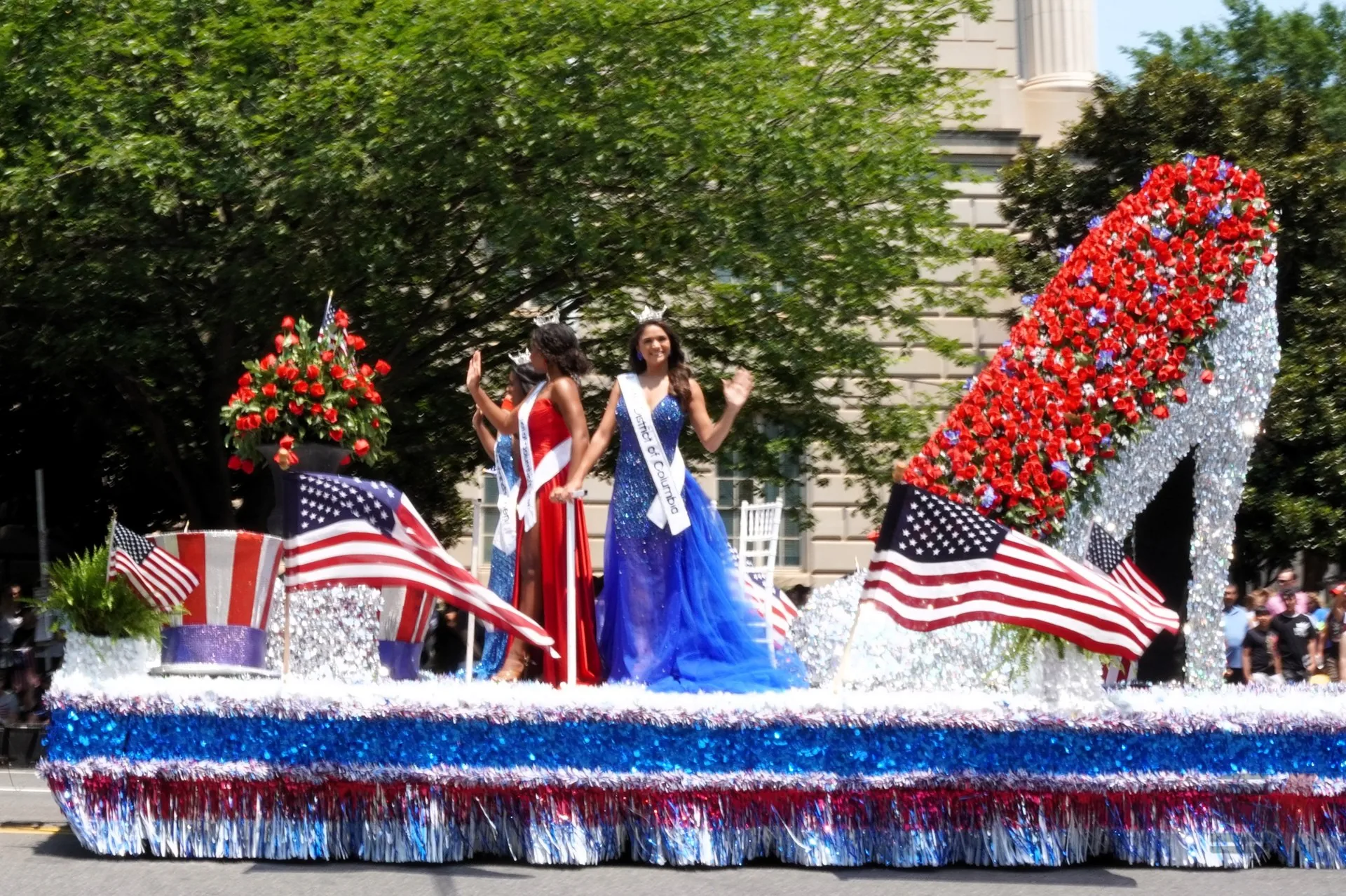 Two contestants in vibrant dresses waving on a float during a parade.