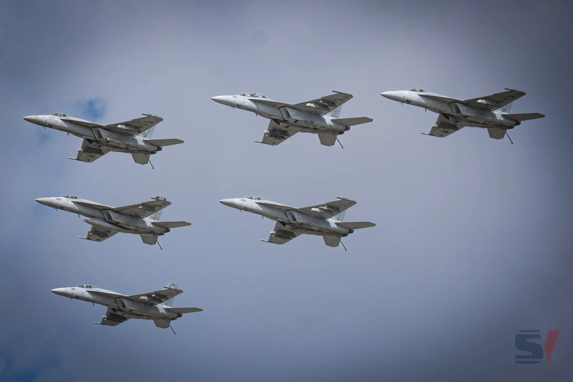 Six F-18 Hornets flying in formation against a cloudy sky.