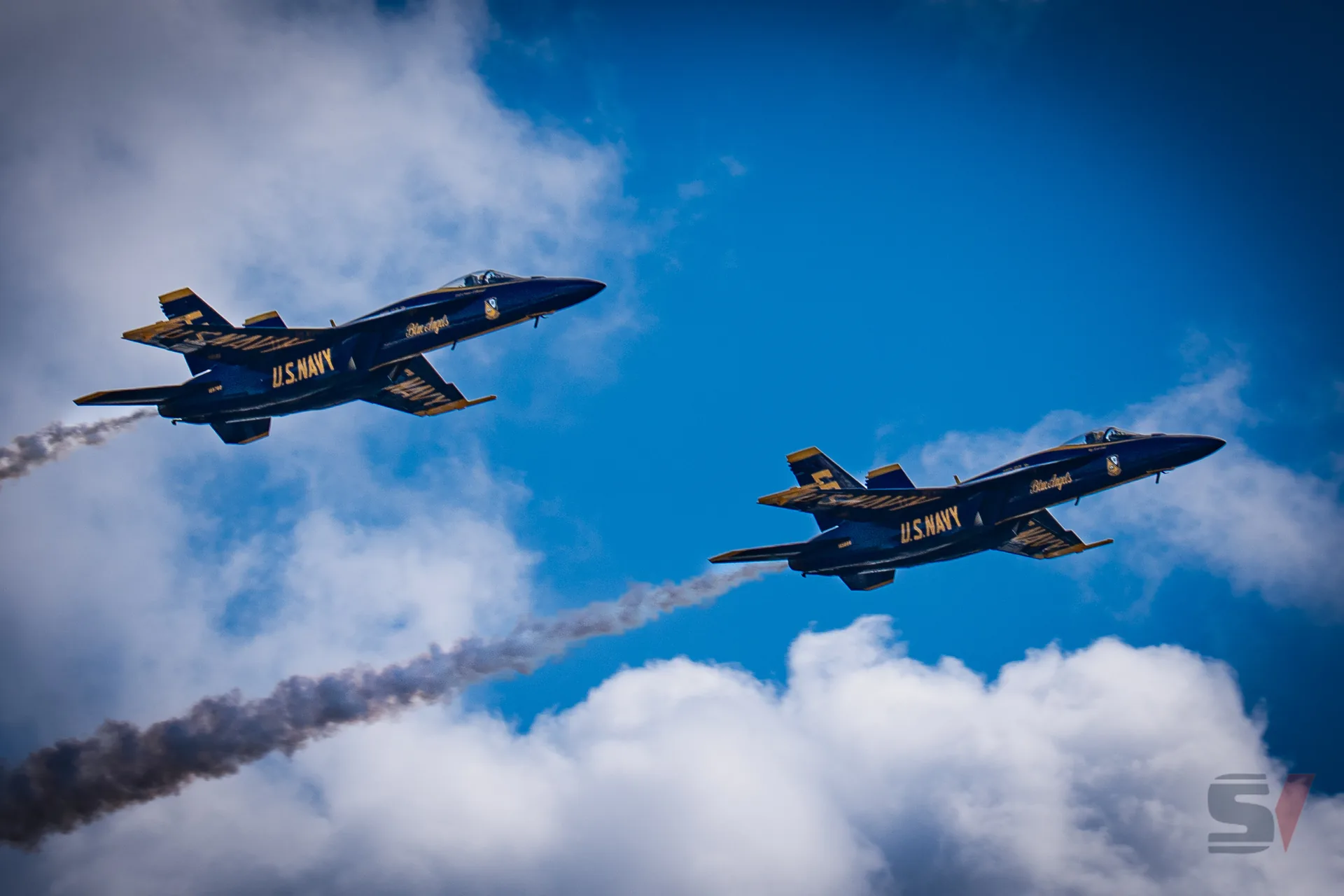 Two Blue Angels jets perform a synchronized aerial maneuver, leaving smoke trails.