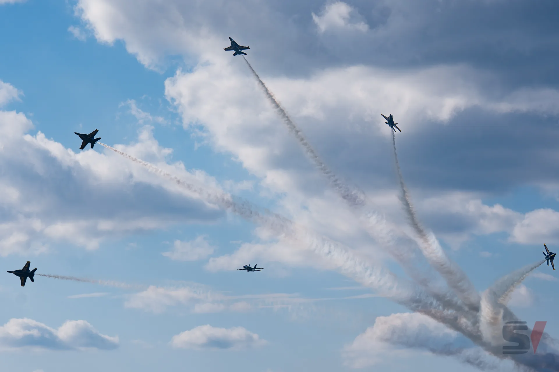 Five Blue Angels jets perform aerial maneuvers with smoke trails in the sky.